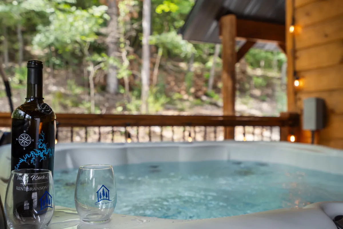 An inviting outdoor hot tub is featured front and center, with a bottle of wine and glass perched on the edge. The background is filled with natural greenery and the warm hues of sunset, capturing a tranquil, relaxing mood ideal for socializing.