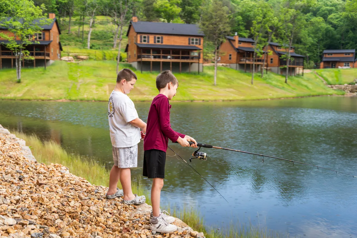 Two boys are fishing on the edge of a calm lake, with wooden cabins visible in the background and lush greenery surrounding the scene. The environment suggests a peaceful, natural setting ideal for outdoor activities.