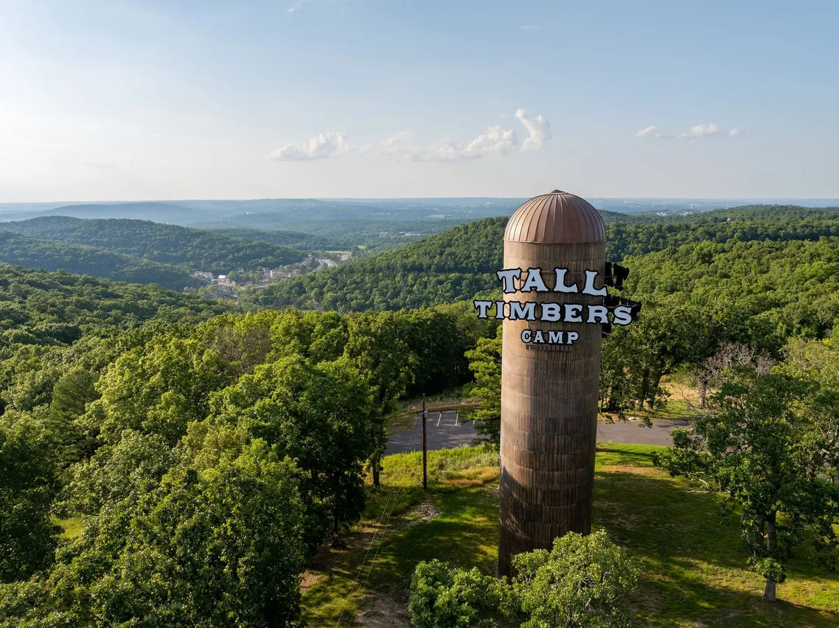 A tall water tower marked 'Tall Timbers Camp' rises above a lush, green landscape with distant hills and valleys visible in the background. The scene is bright and fresh, capturing the expansive outdoor environment and the beginnings of a growing community.