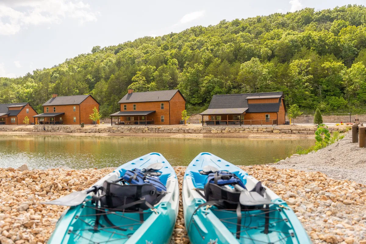 Kayaks rest along the shore in the foreground, with cozy cabins lining the water’s edge and a lush green hillside rising behind. The scene conveys a sense of adventure and tranquility, inviting couples to unwind by the water or embark on a leisurely paddle together.