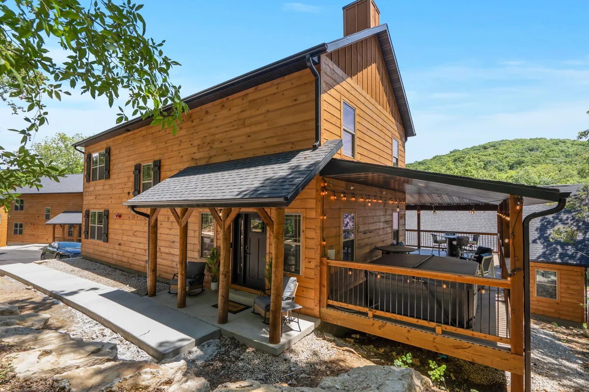 A spacious wooden deck with ample seating is surrounded by trees, offering an inviting space for outdoor dining or evening gatherings. String lights and sunset lighting create a cozy, communal atmosphere.