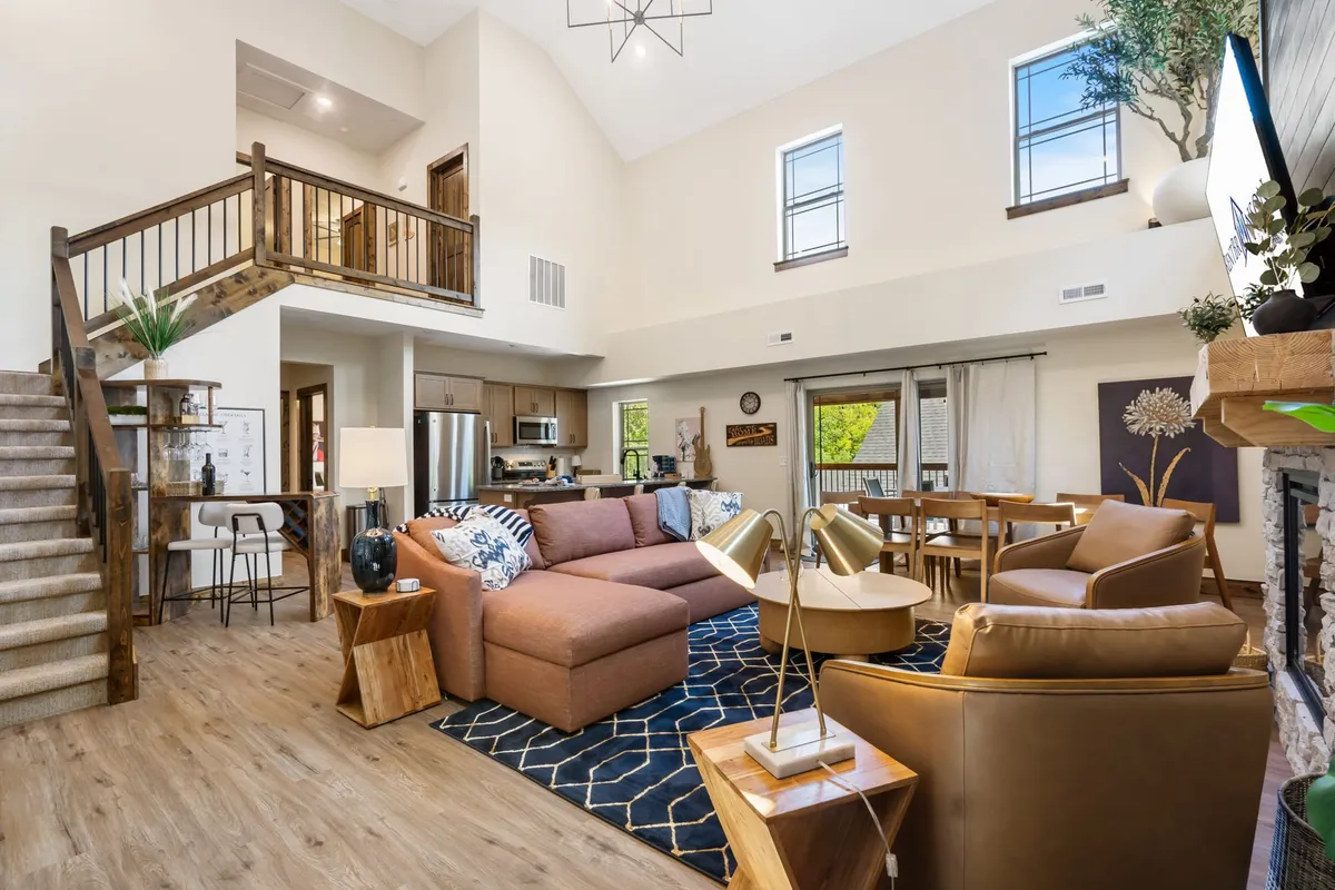 This inviting interior scene features a spacious open-concept layout with a large sectional sofa, a wooden staircase, a dining table, a coffee table with lamps, and a glowing fireplace. The overall ambiance is cozy and welcoming, ideal for family gatherings and relaxed evenings in.