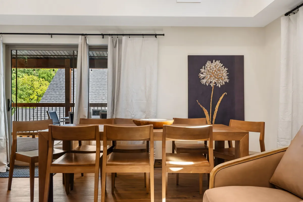 This warm dining area is anchored by a substantial dining table, soft curtains, and a window that brings in natural light. An art piece on the wall adds a touch of personality and charm, while the overall vibe is inviting—perfect for shared meals, board games, or coffee mornings.