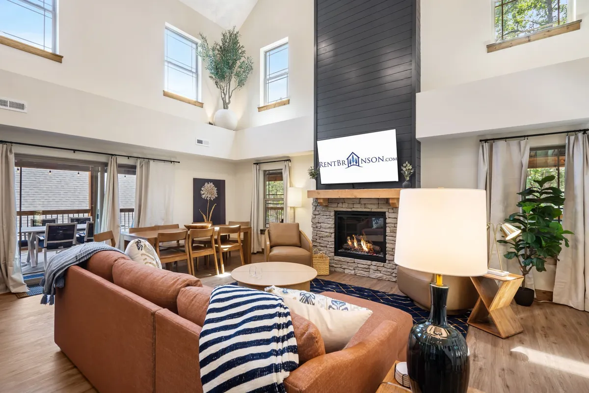 A cozy, sunlit living area featuring a crackling stone fireplace, plush sofa, rustic dining table, and expansive windows that flood the space with natural light. The inviting setup hints at warm family gatherings, relaxed mornings, and tranquil moments overlooking the Ozark woods.