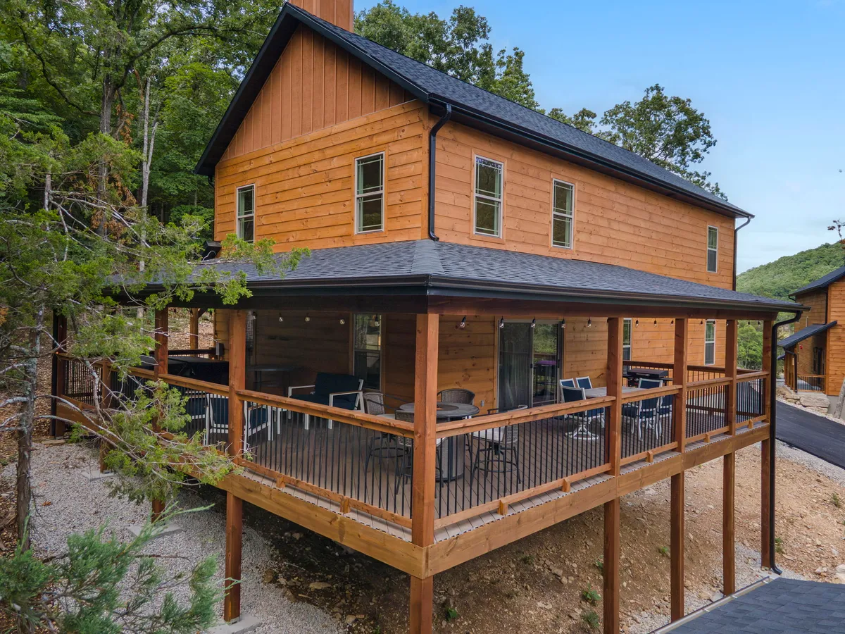 A charming wooden cabin with a wrap-around porch nestled among mature trees. The outdoor seating area invites guests to relax and take in the serene natural surroundings, perfect for enjoying a morning coffee or an evening under the stars. The setting conveys a sense of privacy and coziness—ideal for couples seeking a romantic escape.