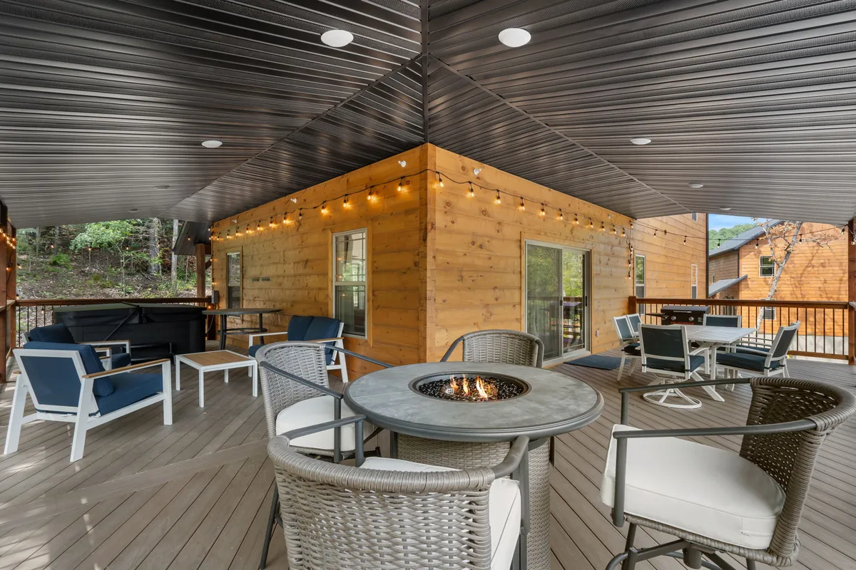 A cozy wooden deck is shown featuring an outdoor fire pit surrounded by comfortable seating and illuminated by warm string lights. The space is designed for socializing and relaxation, with natural surroundings visible beyond the deck.