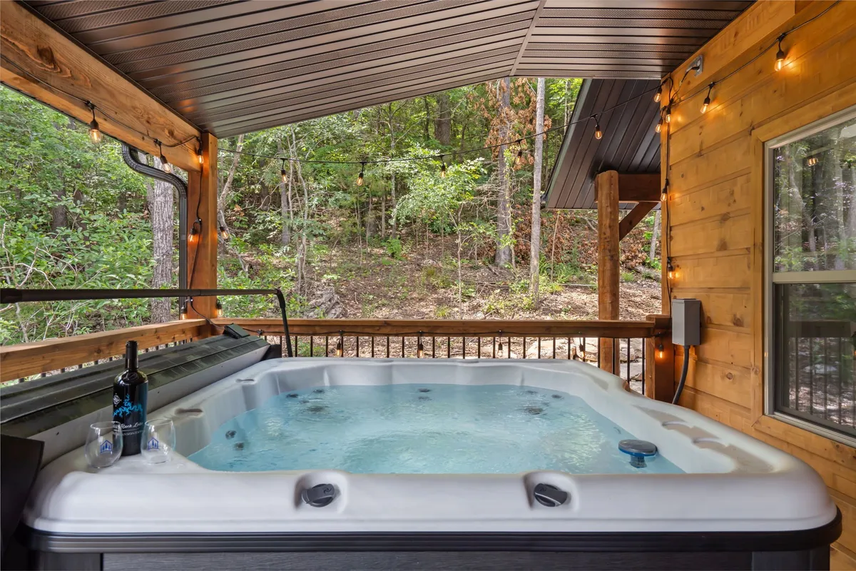 This image features a private hot tub set outdoors with string lights overhead and a bottle of wine on the edge, creating a tranquil and intimate atmosphere. The surrounding deck area is visible, framed by natural greenery, evoking a sense of relaxation under starlit skies.
