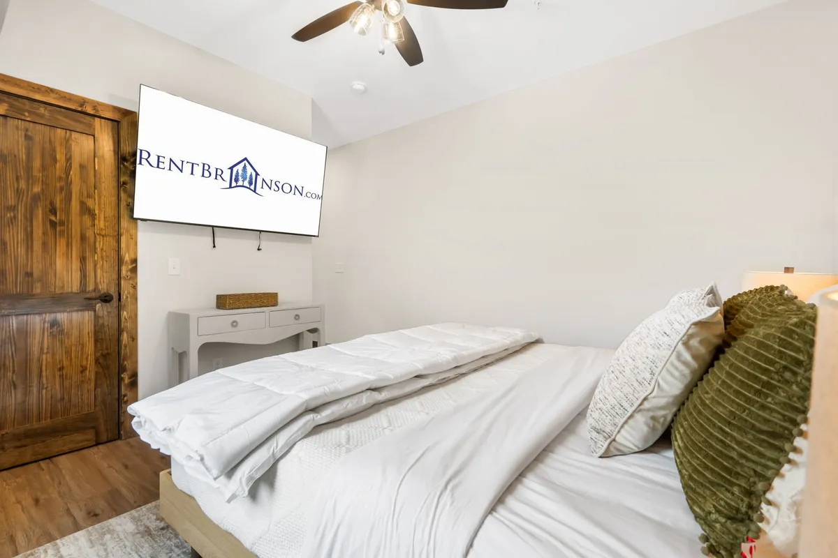 A tranquil bedroom with a large bed dressed in crisp white linens and green accent pillows, a wooden door, and a wall-mounted television. The space is styled in calming earth tones, offering a cozy sanctuary for rest and rejuvenation.