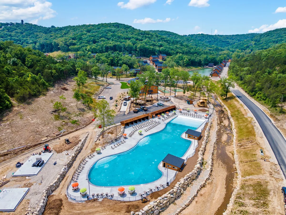 A sparkling outdoor swimming pool sits at the heart of the property, bordered by lounge chairs, cabana structures, and vibrant greenery. A road leads visitors toward the pool, and the scene feels lively and inviting.